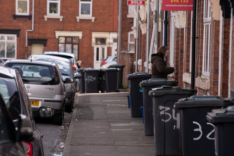 A residential street with a row of dark grey wheelie bins lined up along the pavement in front of a brick building, some with visible white graffiti numbers on their sides. A woman dressed in a dark coat is standing next to one of the bins, seemingly managing household rubbish disposal. Several parked cars of different models and colours are situated on the street, occupying most of the available parking space. The brick building features multiple windows with white frames, and a red sign is partially visible near the entryway, indicating real estate or property sale. The scene is illuminated by natural daylight, with an overcast sky providing soft, diffuse lighting. The arrangement and context suggest a typical setup for private waste collection or on-site rubbish disposal in an urban environment, aligning with services offered by companies specialising in household waste removal in the Queen’s Park area.