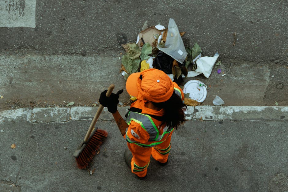 An individual wearing high-visibility orange work clothing with reflective stripes, a matching orange cap, and black gloves is shown from above, using a large push broom with a wooden handle and red bristles to sweep scattered rubbish along a paved sidewalk. The person is positioned near a weathered concrete wall, with a small pile of mixed waste including paper, plastic, and organic material in front of them. The waste appears to be discarded on the ground within a narrow edge, possibly next to a curb or building foundation. Surrounding the scene are typical urban elements such as pavement and concrete, with natural lighting casting soft shadows. This scene exemplifies private waste collection or on-site rubbish clearance, highlighting activities typical of professional rubbish removal services focused on maintaining clean public or private spaces in areas like Queens Park.