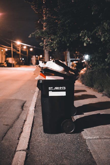 A residential street with a row of dark grey wheelie bins lined up along the pavement in front of a brick building, some with visible white graffiti numbers on their sides. A woman dressed in a dark coat is standing next to one of the bins, seemingly managing household rubbish disposal. Several parked cars of different models and colours are situated on the street, occupying most of the available parking space. The brick building features multiple windows with white frames, and a red sign is partially visible near the entryway, indicating real estate or property sale. The scene is illuminated by natural daylight, with an overcast sky providing soft, diffuse lighting. The arrangement and context suggest a typical setup for private waste collection or on-site rubbish disposal in an urban environment, aligning with services offered by companies specialising in household waste removal in the Queen’s Park area.