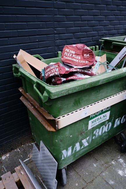 A green wheeled rubbish bin filled with several bags of garden waste, including soil and plant debris, placed on a concrete pavement near a dark brick wall. The bags are red with white text indicating their contents and brand, and some are partially opened. The bin is situated outdoors in an urban setting, with another similar bin visible in the background. The bin's lid is open, exposing the waste inside, and a white plastic container with green text is attached to the side of the bin. The area appears clean and organized, consistent with professional waste collection or rubbish removal services such as those offered by companies like Rubbish Removal Queens Park, providing an alternative to council refuse collection for garden or bulky waste disposal.